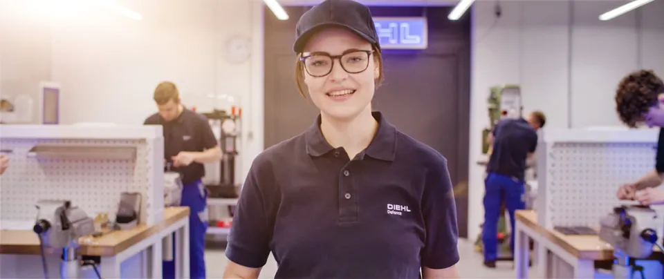 Junge Frau im Diehl Defense Poloshirt lächelt in der Lehrwerkstatt in Nonnweiler, Szene aus dem Azubi Recruitingfilm