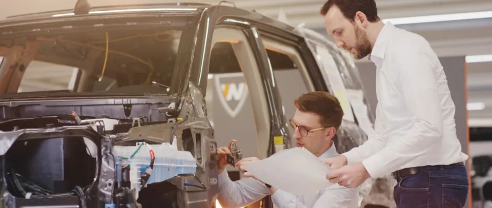 Zwei Ingenieure mit Plänen an einem Fahrzeug im Umbau, Szene aus dem Automotive Imagefilm der Welp Group