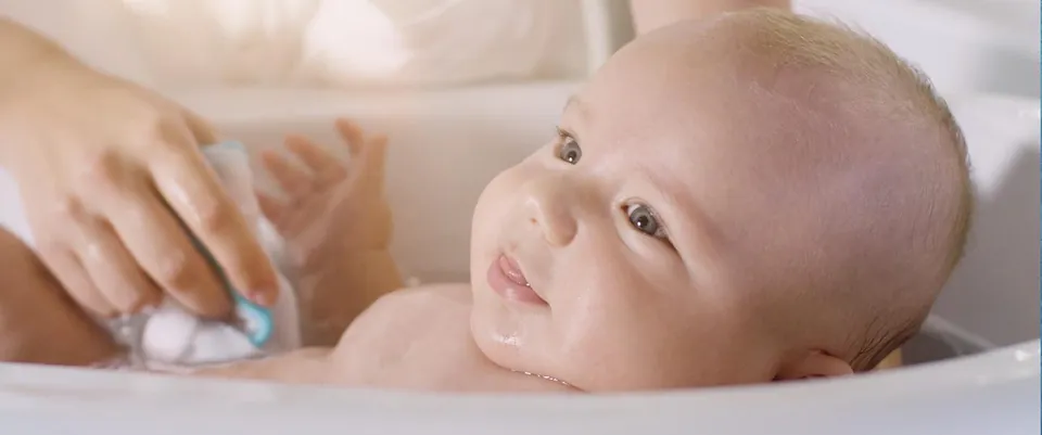 Baby lächelt in der Badewanne, während eine Hand es mit einem Schwamm wäscht, Szene aus dem Produktvideo für Rotho Babydesign